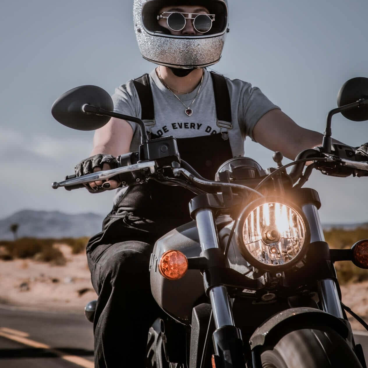 A woman riding a motorcycle wearing Boondocks overalls and a glittery helmet 