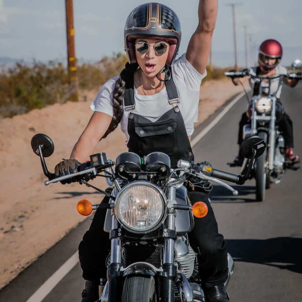 A woman happily riding a motorcycle with one arm raised wearing Boondocks riding overalls