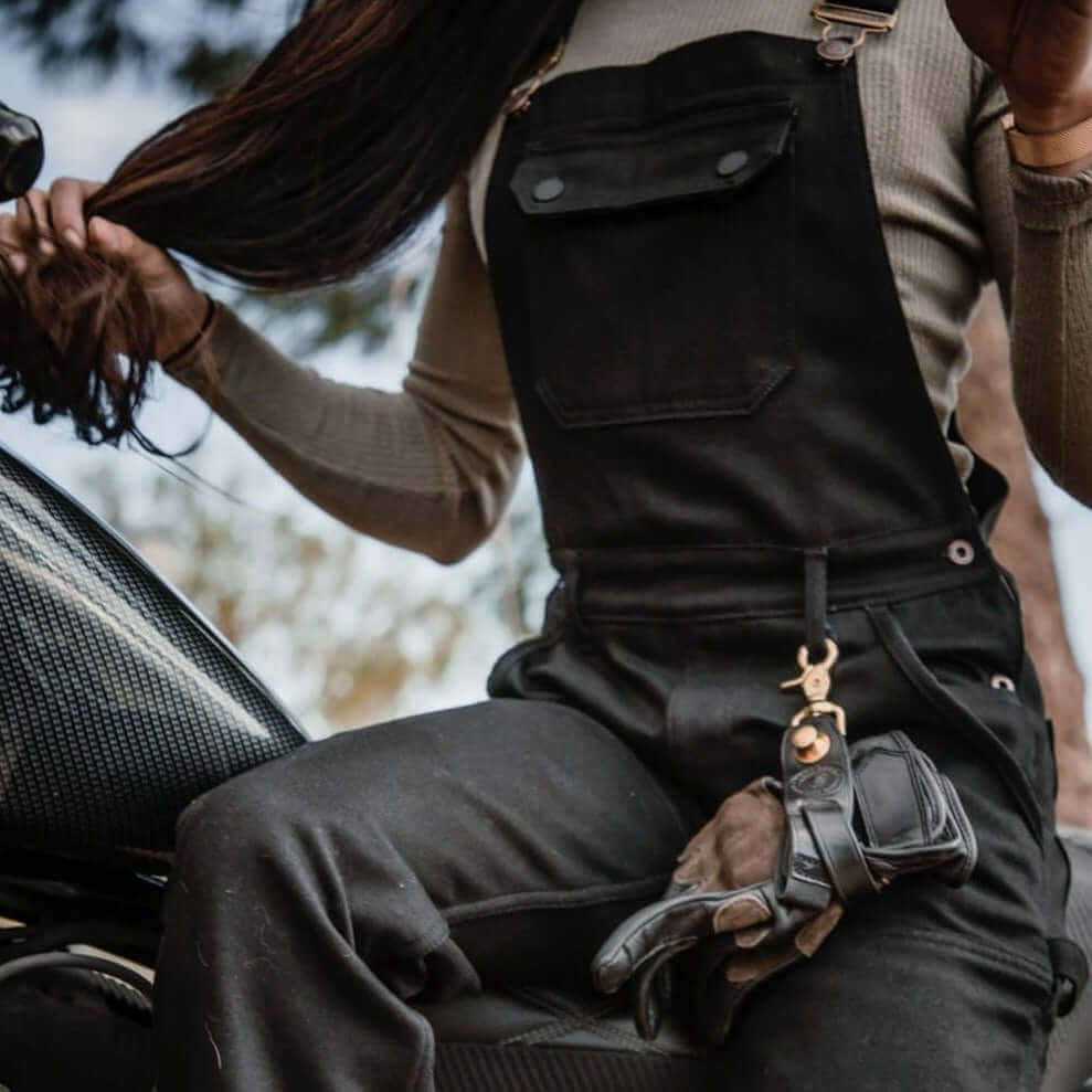 A woman sitting on a motorcycle wearing Tobacco Boondocks overalls with riding gloves clipped to her belt loop